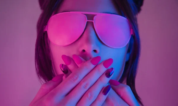 Retrowave style portrait of young woman in pink glasses. Young woman in pink glasses covers her mouth with hands with a manicure, retrowave style portrait. Demonstration of a beautiful manicure.
